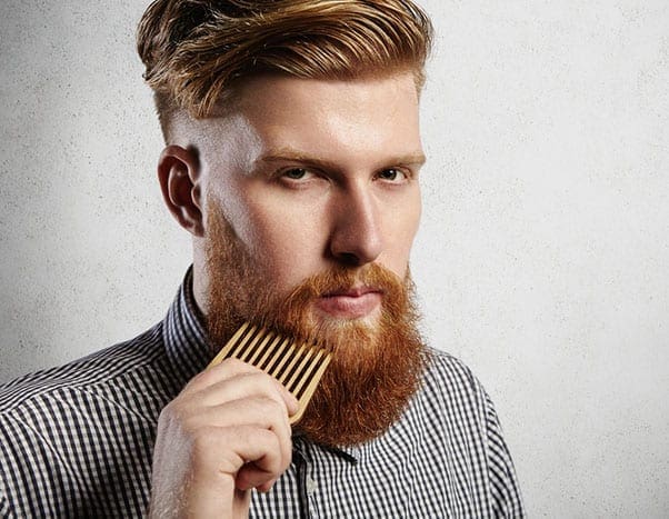Portrait of young attractive redhead hipster male with serious and confident look, holding wooden comb and doing his thick beard. Stylish bearded barber in checkered shirt combing in salon. Horizontal