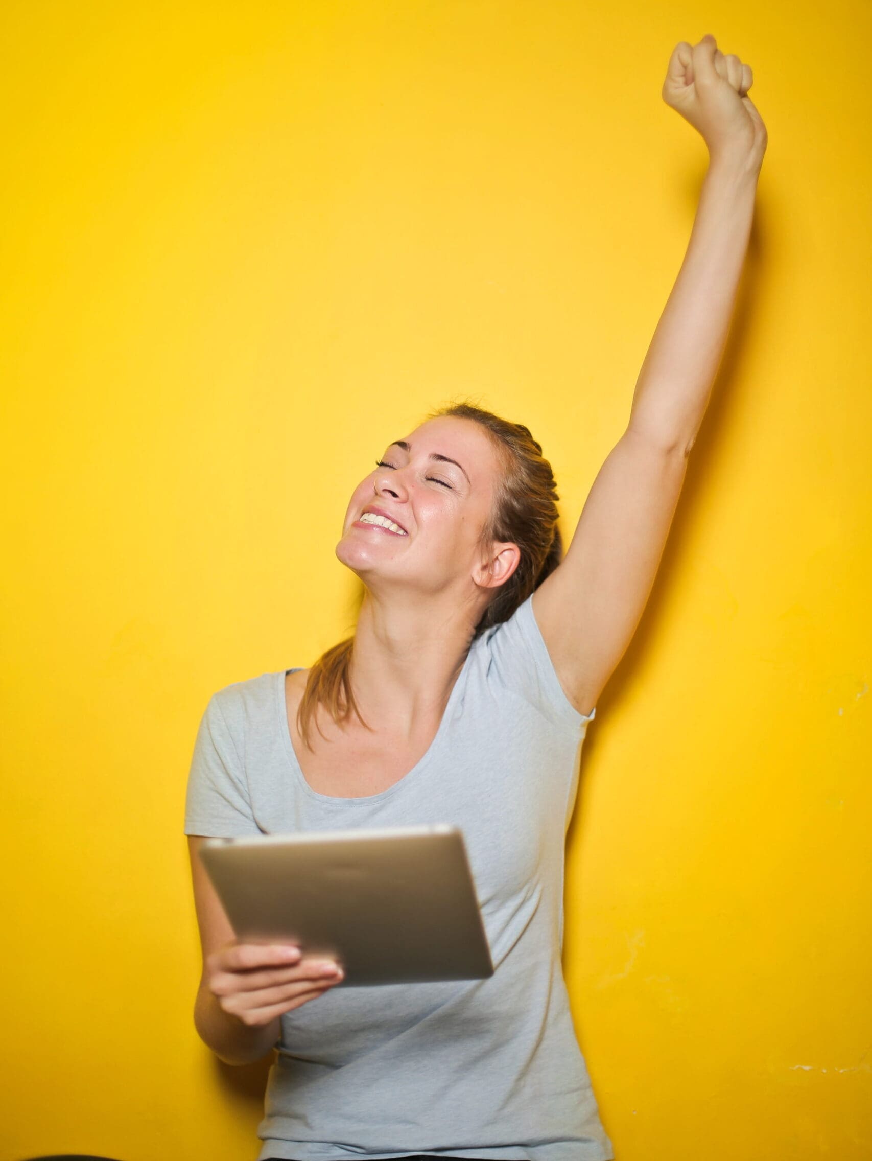 Excited Young Woman Holding An iPad With One Hand Up Against The Yellow Wall